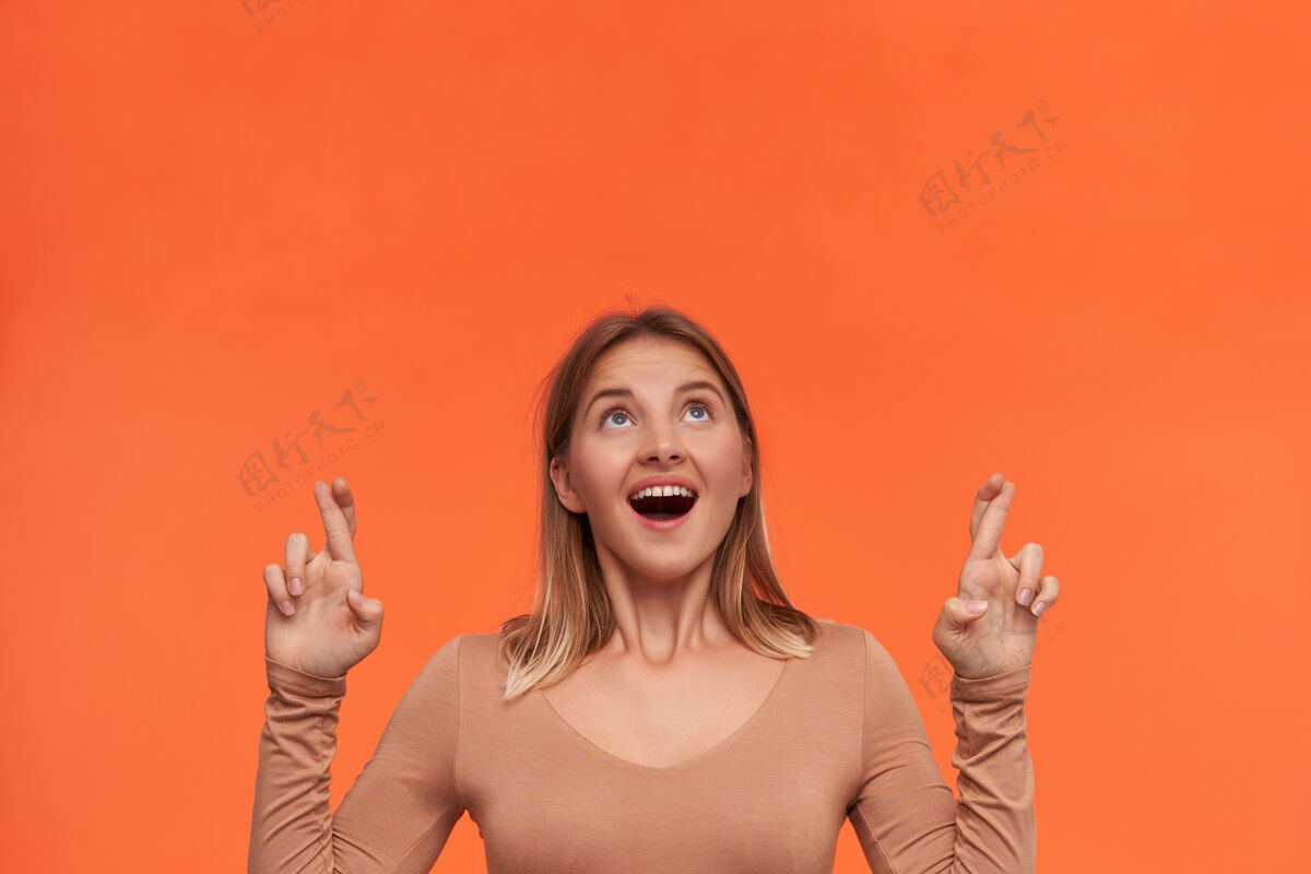 穿激动的年轻迷人的短发金发女郎 身着米色上衣 双手交叉着手指 兴奋地向上看 站在橙色的墙上女士女性提高