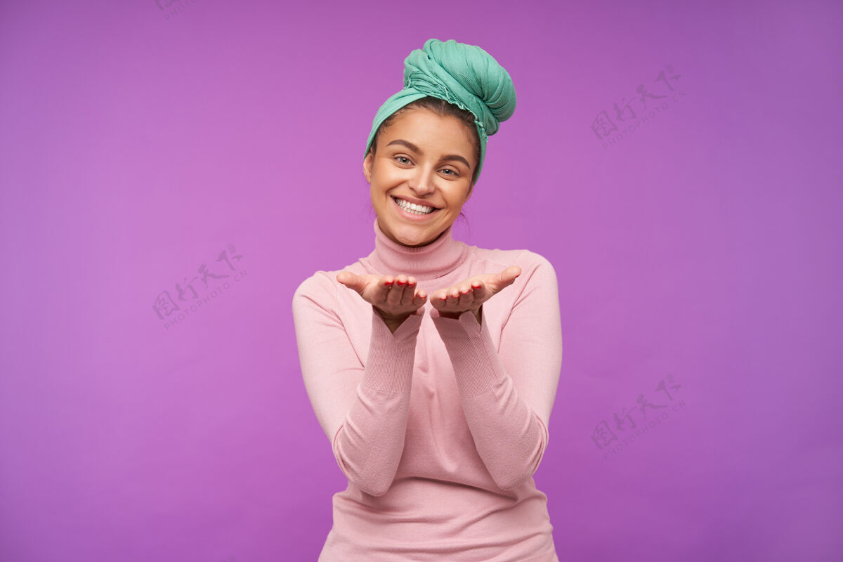 休闲幸福的年轻漂亮的棕色头发的女人薄荷头带保持她的手掌抬起 同时看着前面迷人的微笑 在紫色的墙上摆姿势优雅心情Poloneck