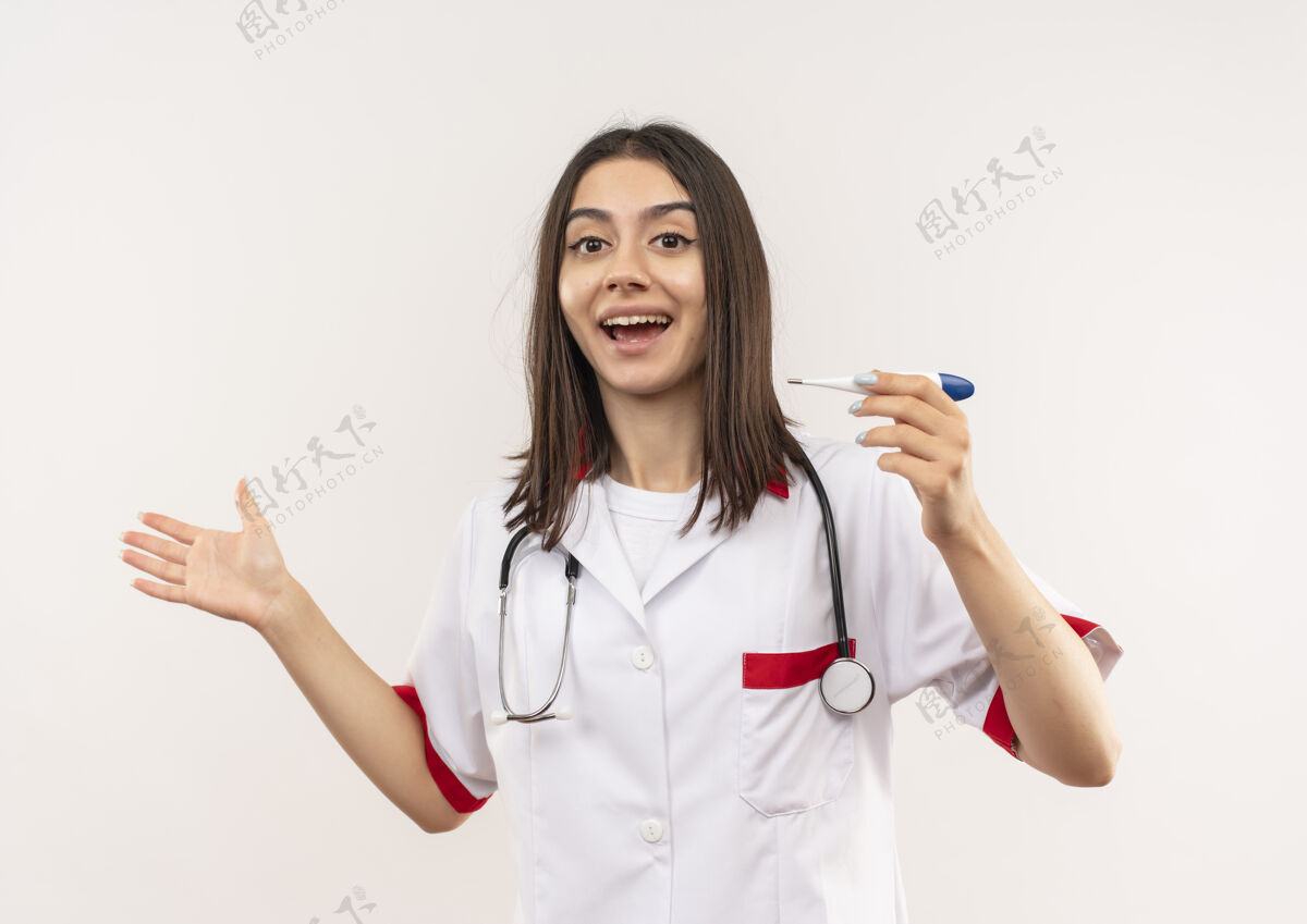 年轻人年轻的女医生穿着白大褂 脖子上戴着听诊器 手里拿着数字体温计 兴奋地用胳膊指着站在白墙上的一边指着市民胳膊