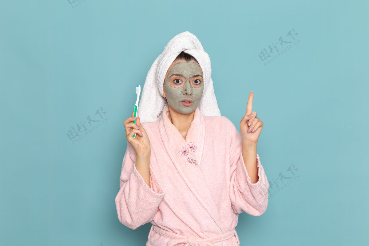 人正面图身着粉色浴袍的年轻女性手持牙刷在蓝色办公桌上清洗美容自护霜淋浴年轻人人视图