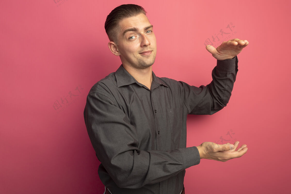 微笑穿着灰色衬衫的年轻帅哥看着前面微笑着自信地展示着手的大小手势测量符号站在粉红色的墙上粉色立场英俊
