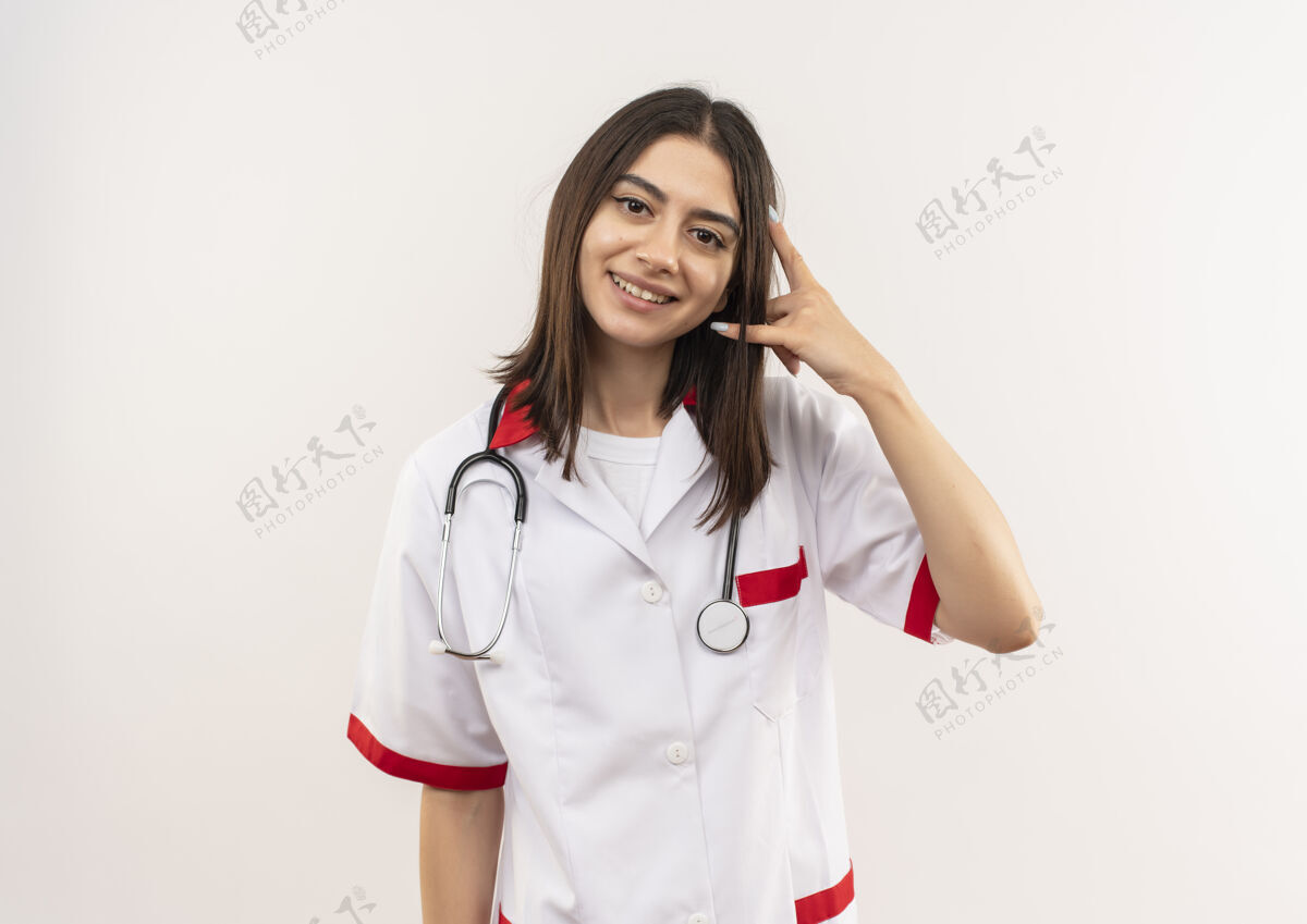 年轻人年轻的女医生穿着白大褂 脖子上戴着听诊器 面带微笑 站在白墙上做着叫我的手势脖子电话手势