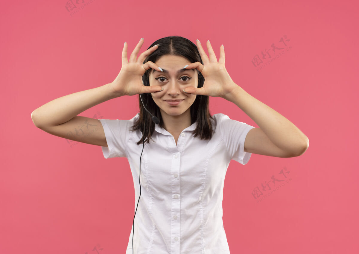 微笑穿着白衬衫戴着耳机的年轻女孩 站在粉红色的墙上微笑着睁开眼睛和手指衬衫公民手势