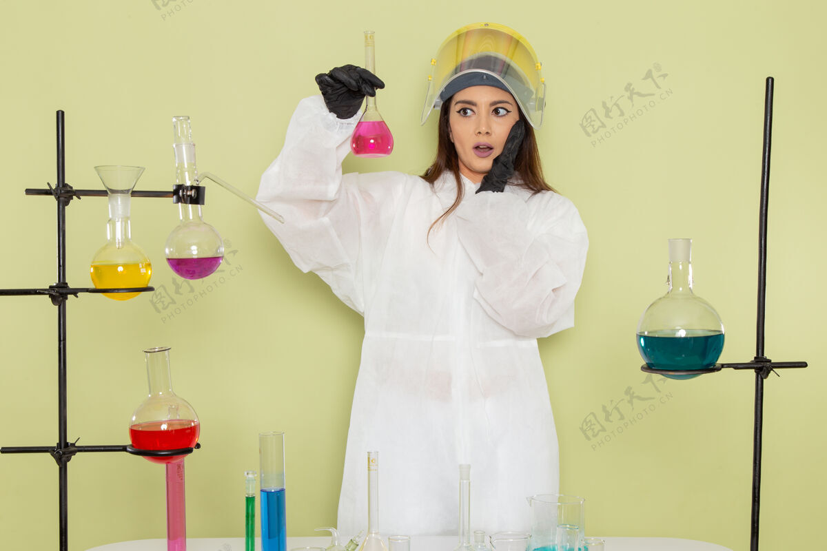 观点正面图身着特殊防护服的年轻女化学家拿着装有溶液的烧瓶在绿色墙上化学工作女科学实验室科学外套女性