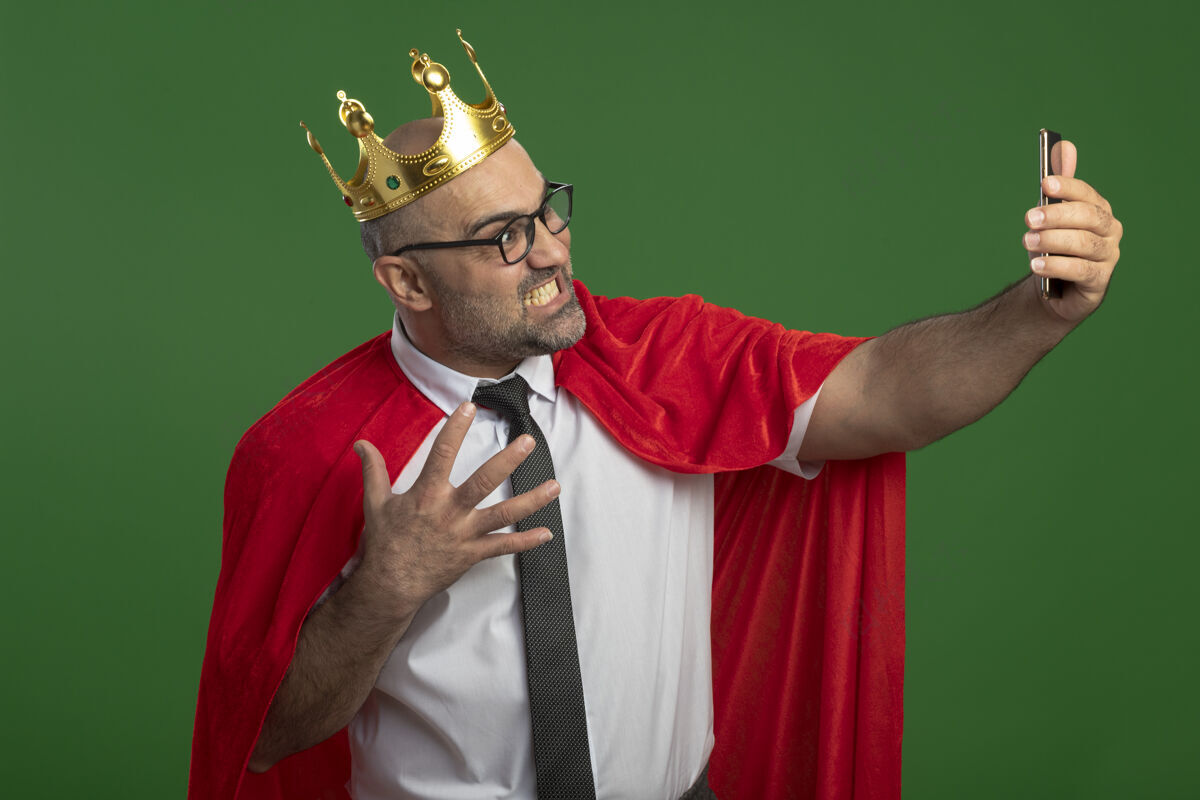 疯狂穿着红斗篷戴着皇冠的超级英雄商人用智能手机自拍疯狂愤怒地站在绿色的墙上国王红色手机