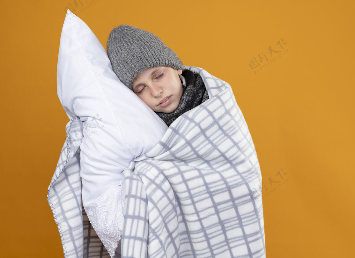 帽子生病的小男孩戴着暖和的帽子 裹着毛毯发烧 抱着枕头站在橙色的墙上抱着小穿