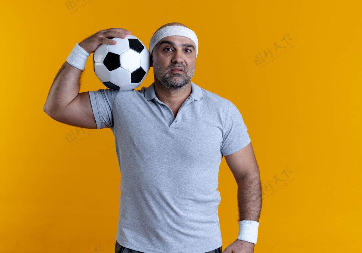 教练戴着头巾的成熟运动型男子举着足球望着前方 严肃的脸站在橙色的墙上头带成熟严肃