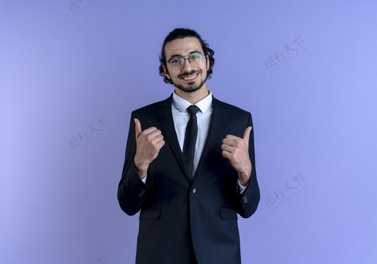 西装一位身穿黑色西装 戴着眼镜的商人站在蓝色的墙上 面带微笑 兴高采烈地竖起大拇指男人公司男人