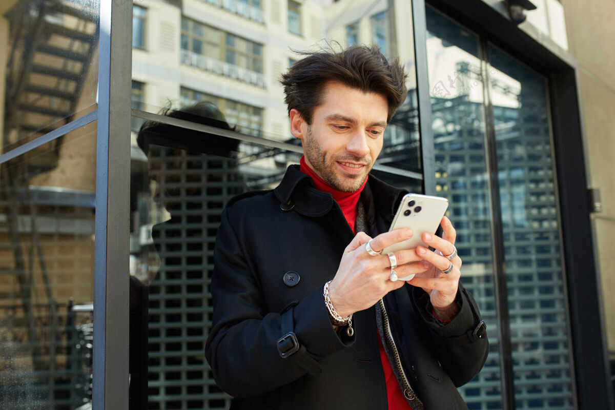 头发一张棕色头发的年轻人举着手机 站在城市背景上积极地看着屏幕的户外照片风衣手帅哥