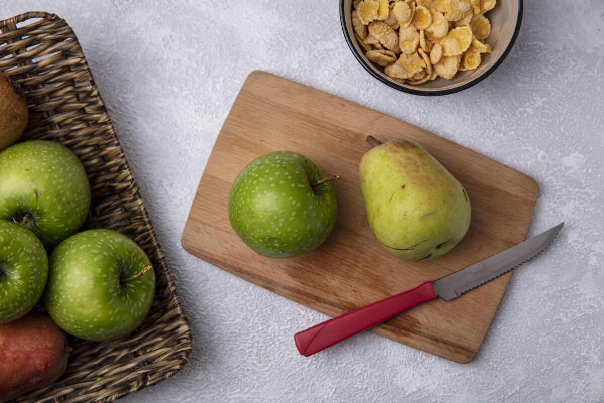 切俯视图绿色的苹果 梨和刀放在砧板上 碗里放着玉米片 背景是白色的白色视图食物