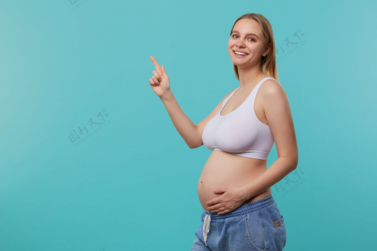 休闲摄影棚拍摄的年轻漂亮的白头未来妈妈手放在肚子上 用食指愉快地向上展示 站在蓝色背景上站着白头喜悦