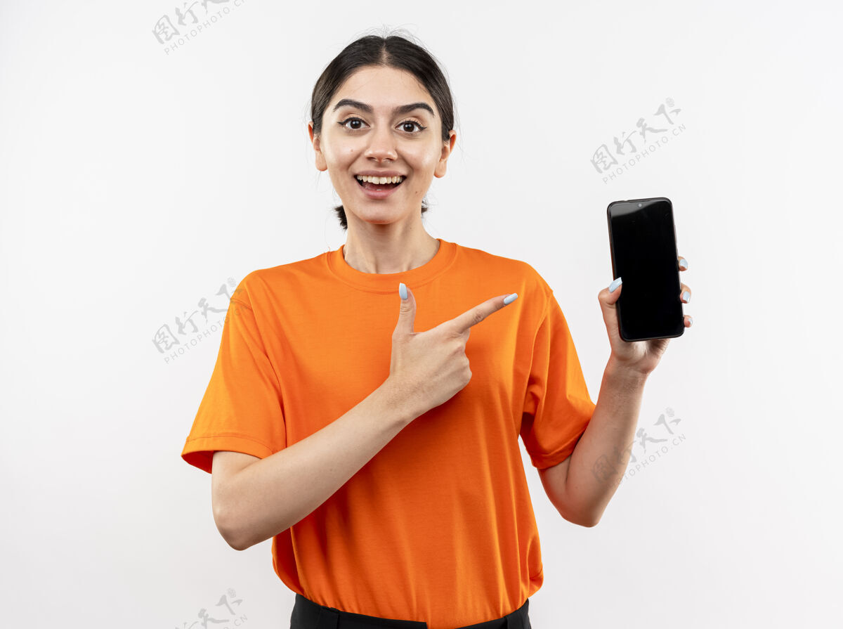 穿着穿着橙色t恤的年轻女孩站在白色的墙上 用食指指着智能手机 开心地笑着橙色年轻目录