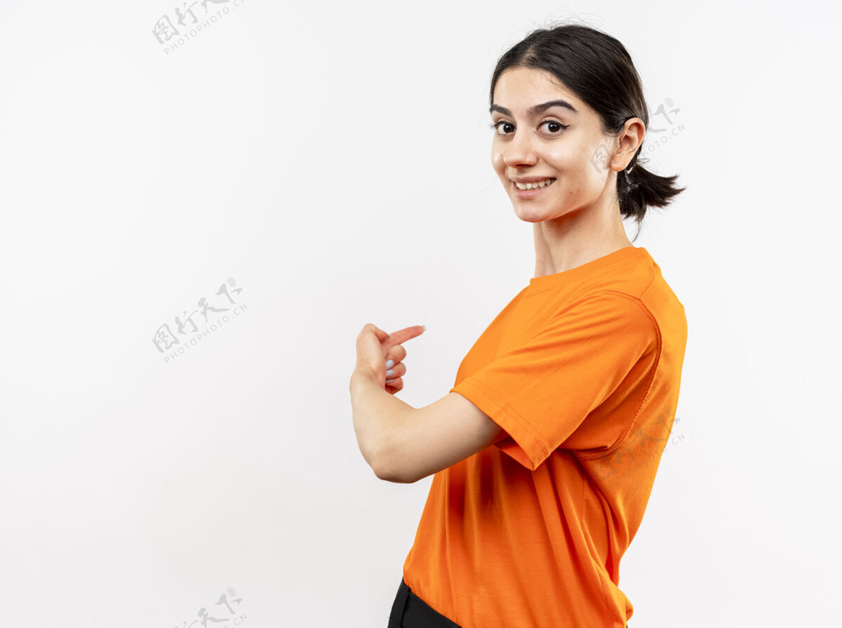 站年轻女孩穿着橙色t恤 脸上带着微笑 手指指向后面 站在白色的墙上穿指指点点背