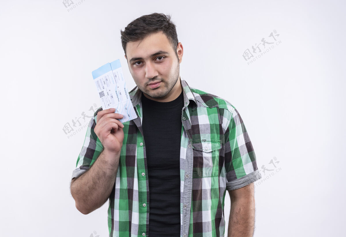 年轻人穿着格子衬衫的年轻旅行者站在白墙上 脸上带着自信的微笑 手里拿着机票旅行者人机票