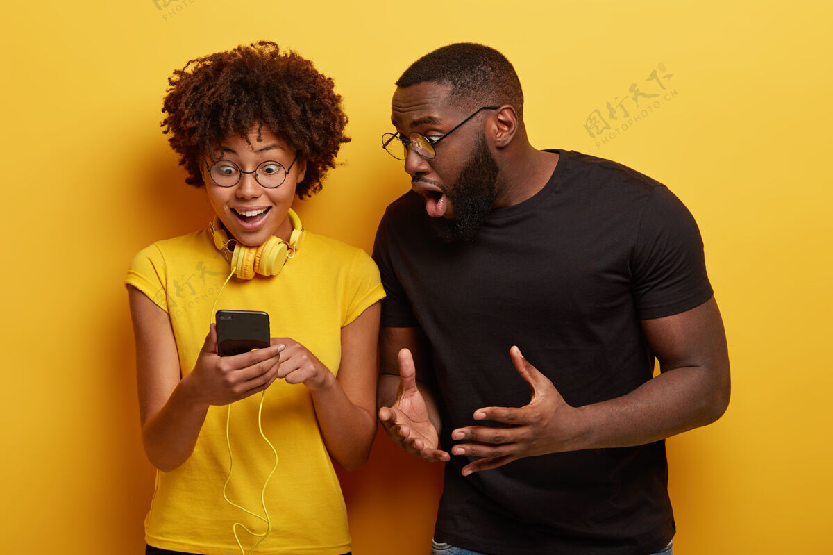 满意高兴的非洲女人盯着智能手机设备连接到耳机男性小玩意聆听