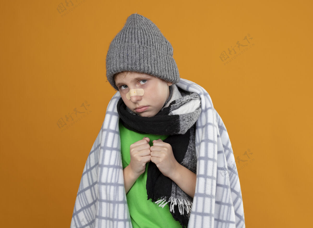 橙色生病的小男孩戴着暖和的帽子 戴着围巾 裹着毛毯 鼻子上贴着补丁 站在橘色的墙上 闷闷不乐 一副病态的样子鼻子生病男孩