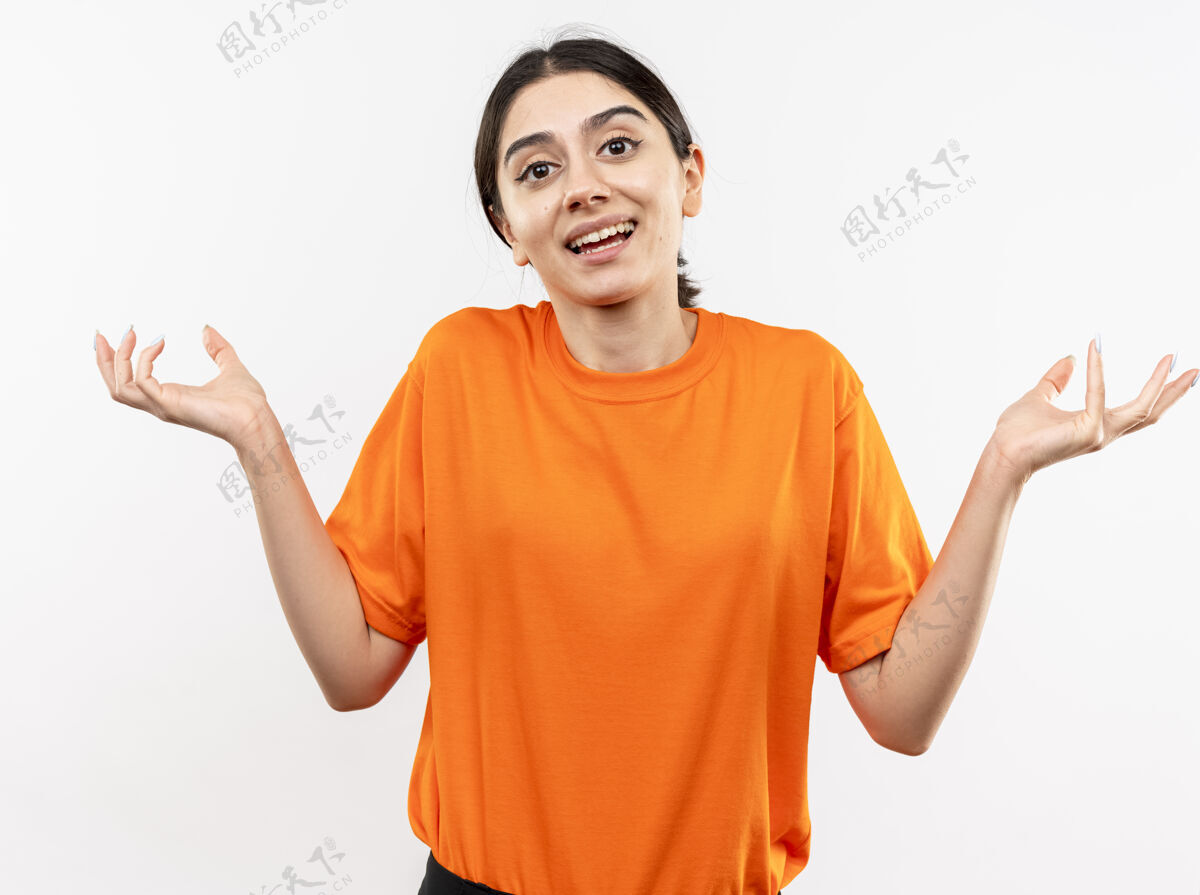 蔓延身穿橙色t恤的年轻女孩困惑而不确定地微笑着张开双臂站在白色的墙壁上疑惑女孩橙色怀疑