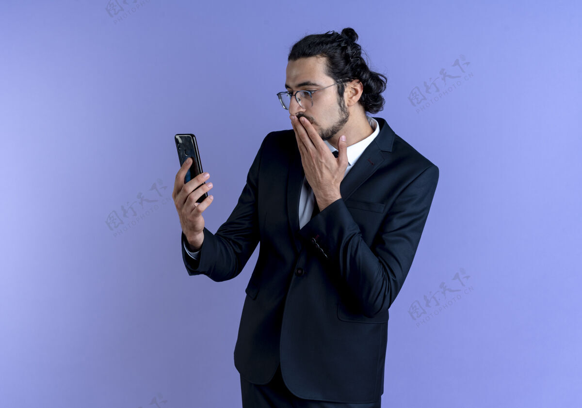 优雅一个穿着黑西装戴着眼镜的商人站在蓝色的墙上 看着智能手机的屏幕 既惊讶又困惑惊喜家伙眼镜