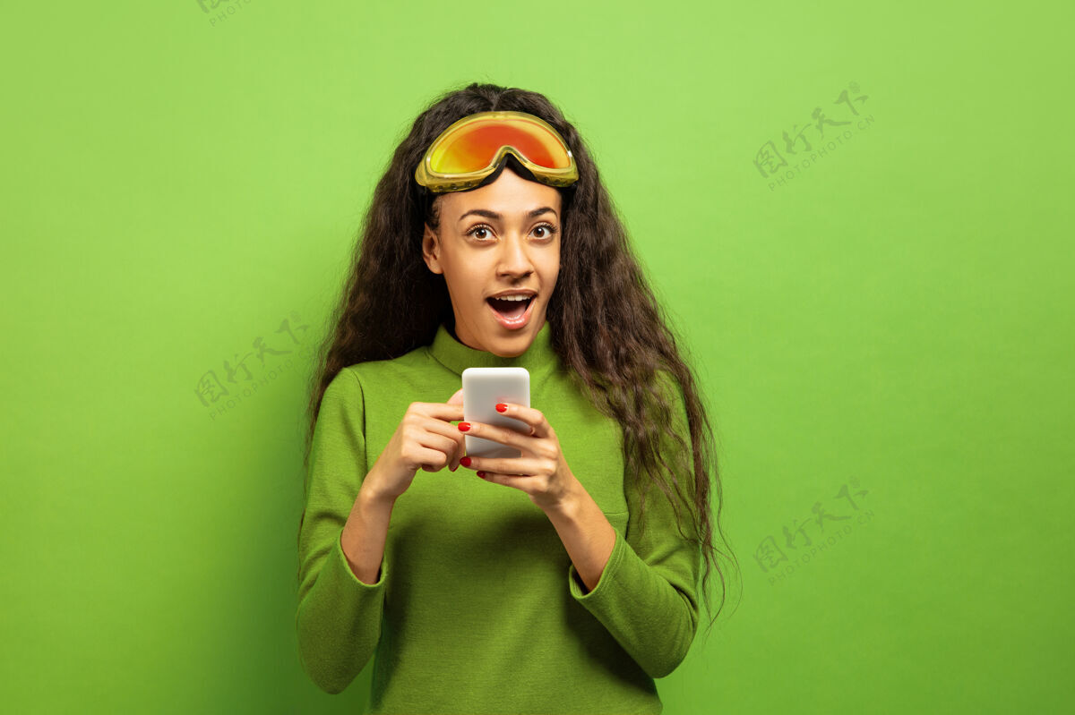 肖像以绿色工作室为背景 黑色皮肤的非洲裔年轻女子的滑雪面具肖像人类情感 面部表情 销售 广告 冬季运动和假期的概念使用智能手机女孩滑雪头发