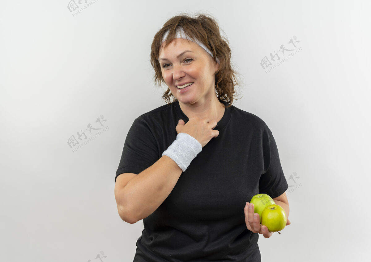 抱着身穿黑色t恤 头箍 手持两个绿色苹果 站在白色墙壁上欢快微笑的中年运动女性市民姿势运动员