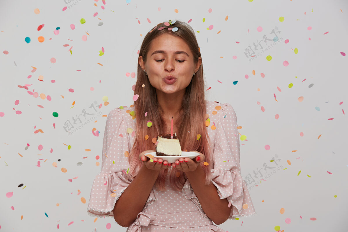 女性室内拍摄的可爱的年轻长发金发女人吹生日蛋糕上的蜡烛 而站在白色的墙壁 穿着粉红色的浪漫礼服金发发型站着