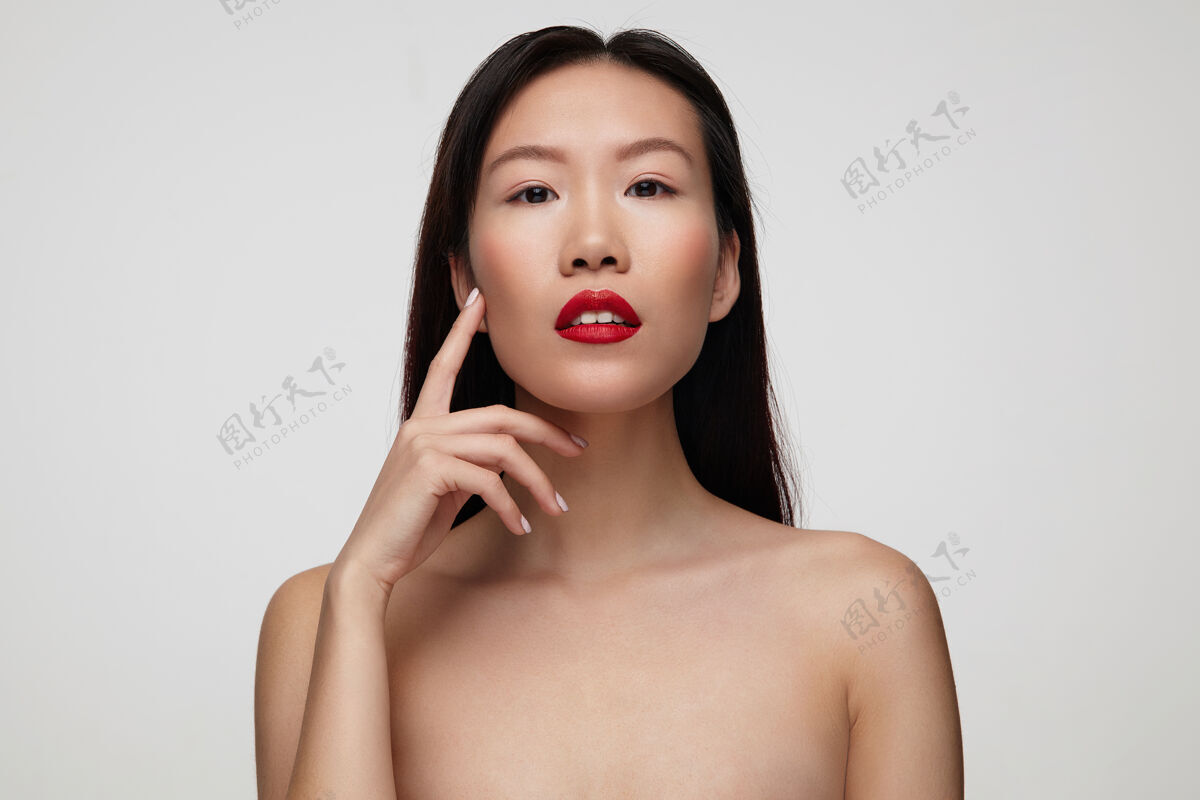 女士平静的年轻漂亮的黑发女人 红唇 把食指举在脸颊上 隔着白墙女性提高头发