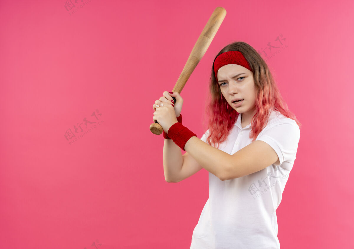 头带戴着头巾打棒球的年轻运动型女子手持球棒 站在粉红色的墙上 看上去很自信人人运动