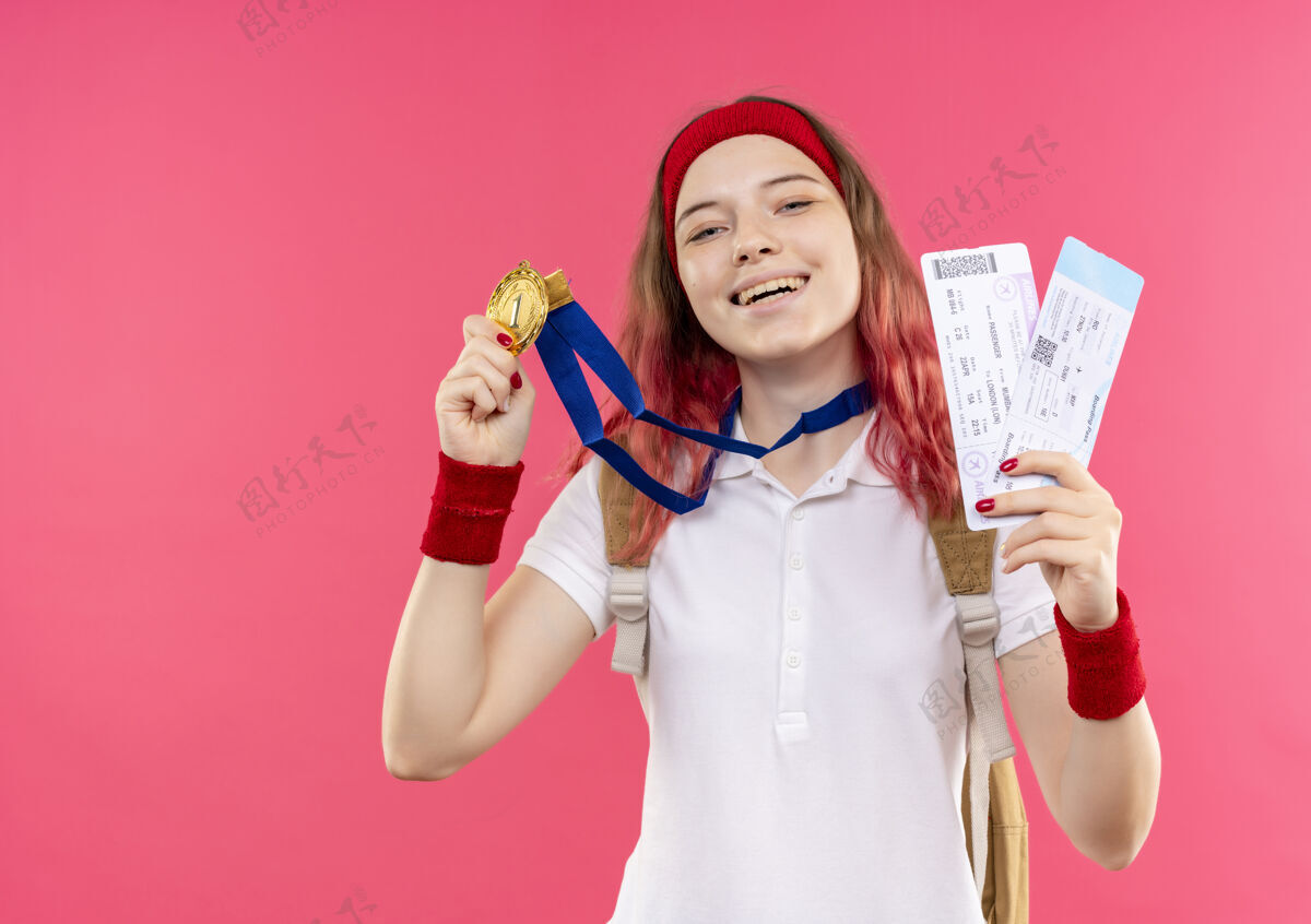 运动装戴着头巾展示金牌的年轻运动女性手持两张机票微笑着站在粉红色的墙上 脸上洋溢着幸福表演微笑脸
