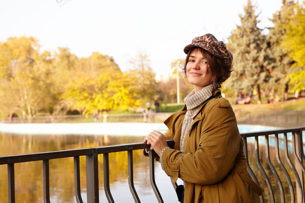 女性积极的年轻迷人的短发黑发女性斜靠在铁栏杆上 在城市公园的湖边摆姿势 愉快地看着 温柔地微笑着树欢呼休闲