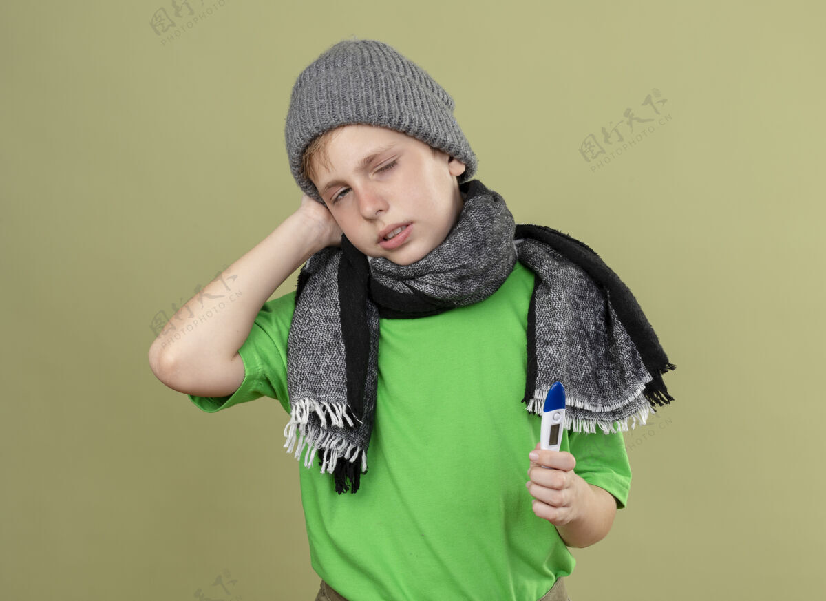抱着生病的小男孩穿着绿色t恤 戴着暖和的围巾和帽子 感觉不舒服 拿着温度计看着相机 站在灯光背景下 不开心 不舒服生病帽子站着