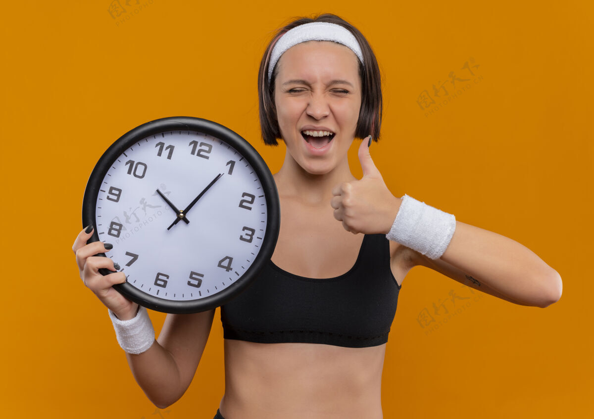 女人身穿运动服的年轻健身女士 头箍举着挂钟 高兴而兴奋地竖起大拇指站在橙色的墙上市民头带运动