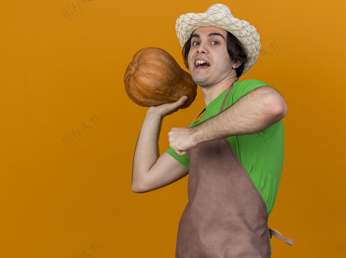 站年轻的园丁 围裙戴帽 手里拿着南瓜 站在橘黄色的背景上 高兴地抛来抛去扔南瓜抱
