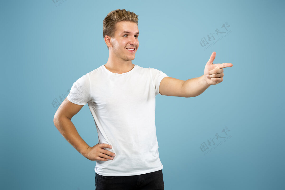 公司蓝色工作室背景上的白人年轻人半身肖像穿着衬衫的漂亮男模人类情感的概念 面部表情 销售 广告指着侧面 微笑人事业手势