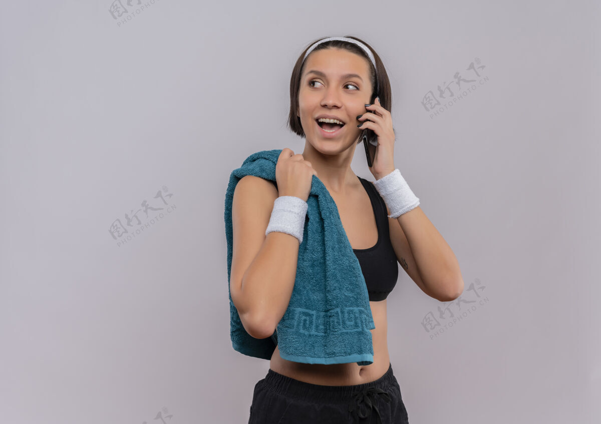 站立身穿运动服 肩上扛着毛巾的年轻健身女士站在白色的墙上 一边高高兴兴地说着手机运动员女人运动