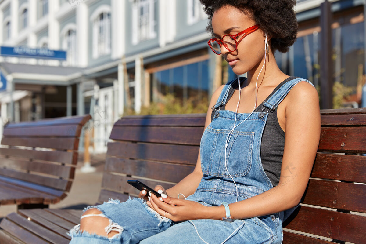 应用程序黑皮肤的时髦女孩的照片 非洲发型 在社交网络中与追随者聊天 用耳机听喜爱的音乐 在户外消遣 坐在木凳上 等待朋友真实放松工作服