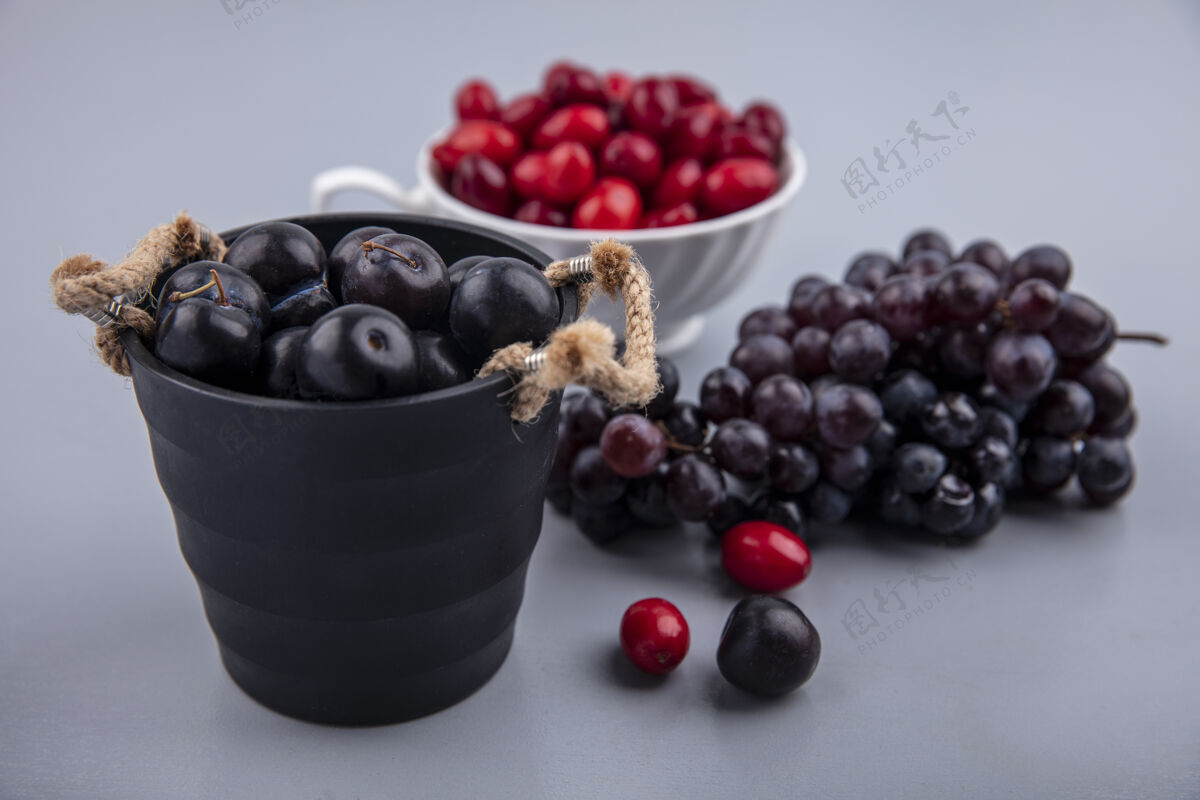 风景在一个黑色的篮子里看到深紫色的黑刺水果 杯子里放着山茱萸 灰色的背景上有葡萄甜味篮子木头