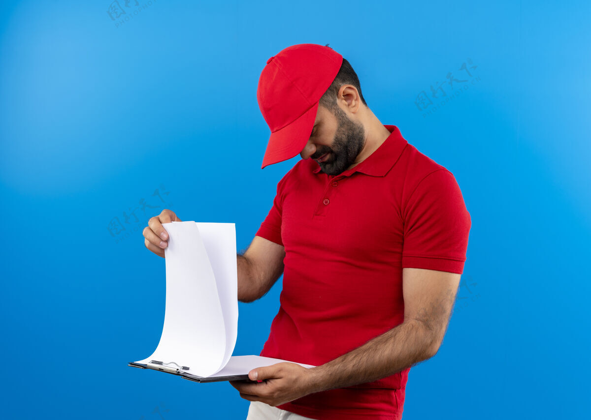 递送留着胡子的送货员穿着红色制服 戴着帽子 手里拿着剪贴板 手里拿着空白页 严肃地站在蓝色的墙上看着他们剪贴板帽子男人