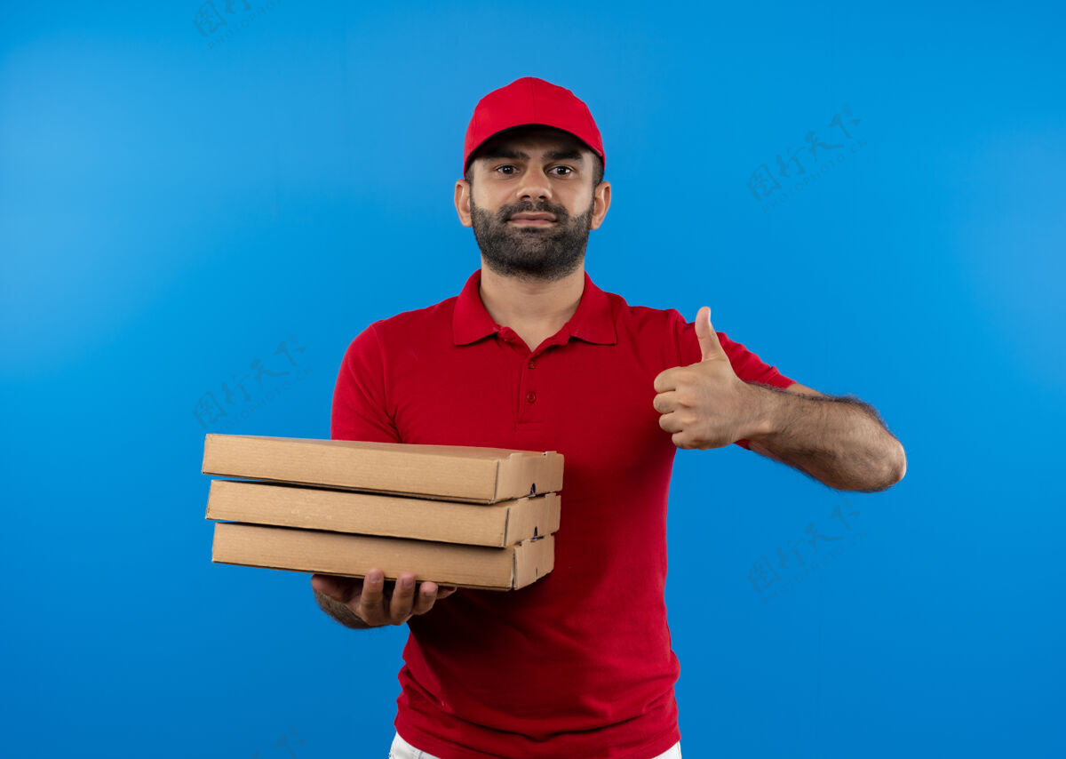 制服满脸胡须 身穿红色制服 头戴鸭舌帽的送货员站在蓝色的墙上 手里拿着一叠披萨盒 竖起大拇指 微笑着自信地站着男人送货帽子