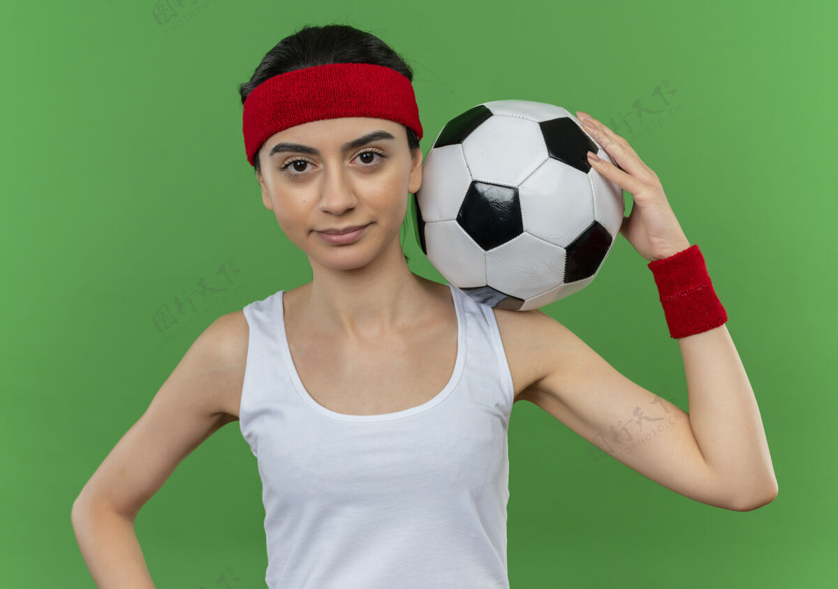 自信身穿运动服 头箍 手持足球 表情自信的年轻健身女士站在绿色的墙上足球头带人