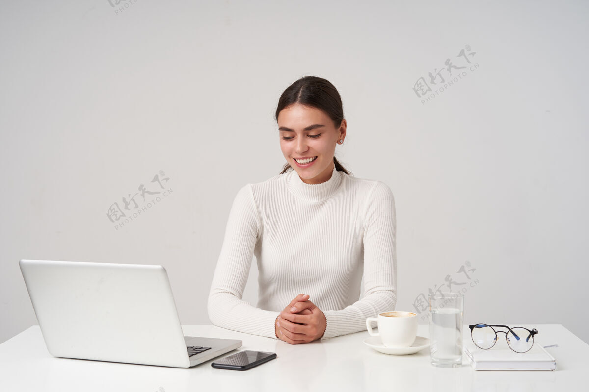 微笑年轻可爱的黑发女士 面容自然 双手合十 坐在白色墙壁上的桌子旁 面带微笑 穿着正式的衣服年轻手机模特