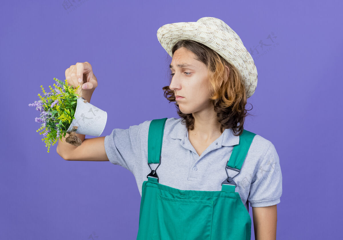 连身衣年轻的园丁穿着连体衣戴着帽子拿着盆栽植物看着它伤心抱表情年轻