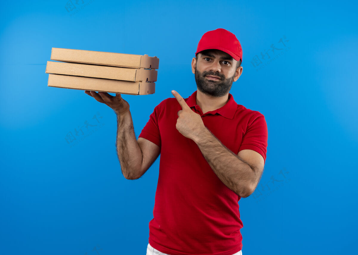 帽子满脸胡须 身穿红色制服 头戴鸭舌帽的送货员拿着一叠披萨盒 用手指指着他们 站在蓝色的墙上自信地微笑着制服自信披萨