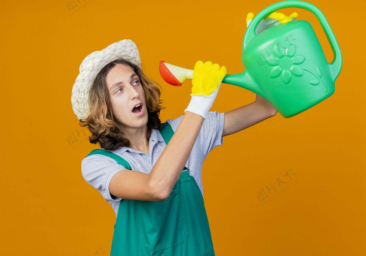 抱年轻的园丁戴着橡胶手套 穿着连体衣 戴着帽子 手里拿着浇水罐花园橡胶帽子