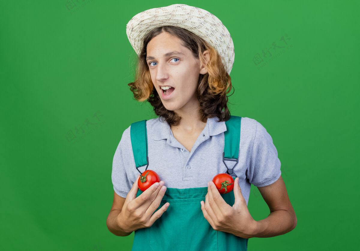 新鲜有趣的年轻园丁 穿着连体衣 戴着帽子 手里拿着新鲜的西红柿微笑连身衣有趣