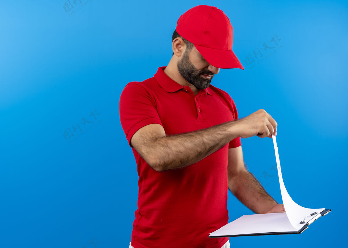 空白留着胡子的送货员穿着红色制服 戴着帽子 手里拿着剪贴板 手里拿着空白页 严肃地站在蓝色的墙上看着他们页面严肃剪贴板