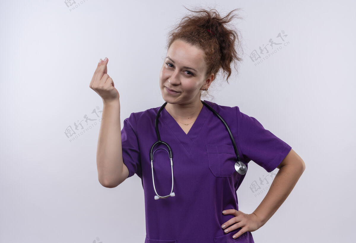 手势年轻的女医生穿着医疗制服 用听诊器搓手指赚钱 脸上带着微笑站在白墙上微笑医生年轻人