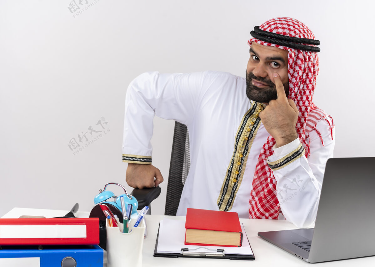 笔记本电脑身着传统服装的阿拉伯商人坐在桌子旁 用手指指着笔记本电脑的眼睛 在办公室里等待付款传统付款手指