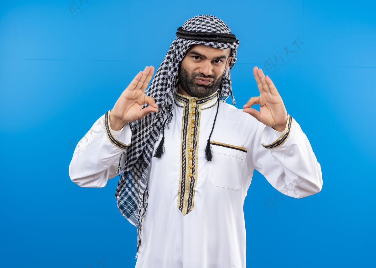 穿一个穿着传统服装的阿拉伯男人站在蓝色的墙上 双手看起来很自信两者看信心