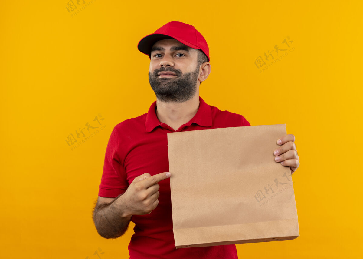 看着一个留着胡须 身穿红色制服 戴着帽子的送货员站在橙色的墙上 用手指着纸袋 带着自信的微笑送货自信胡子