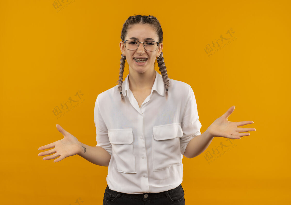 手臂戴着眼镜 梳着辫子 穿着白衬衫的年轻女学生望着前面 微笑着张开双臂站在橙色的墙上姿势微笑衬衫